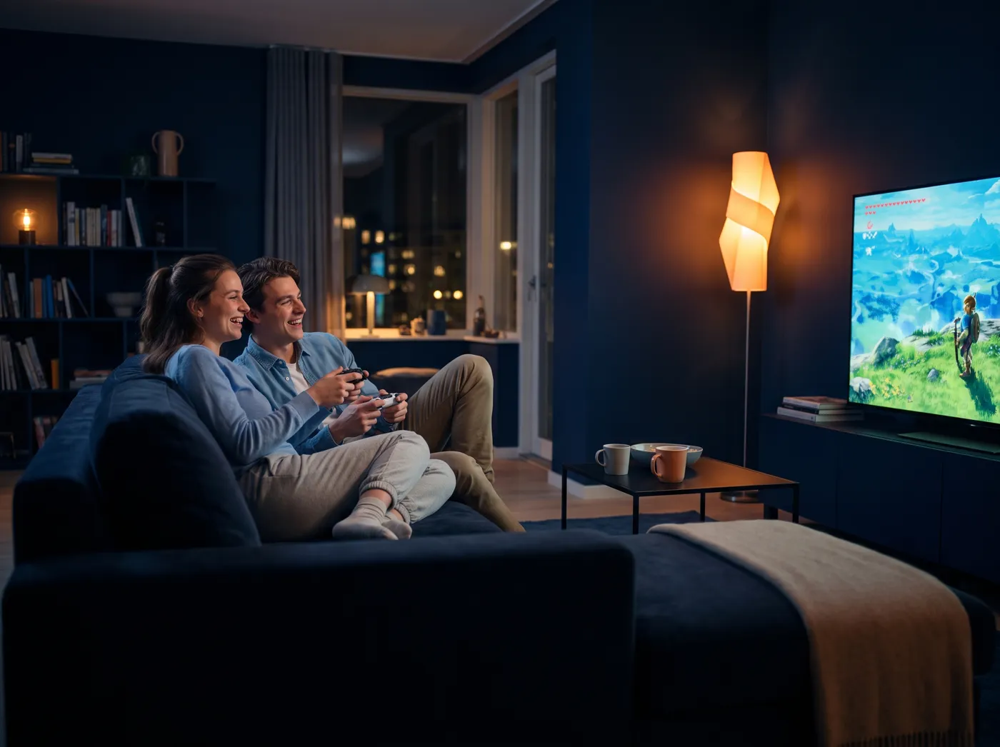 Two friends laughing on a couch, playing an adventure game on a big TV